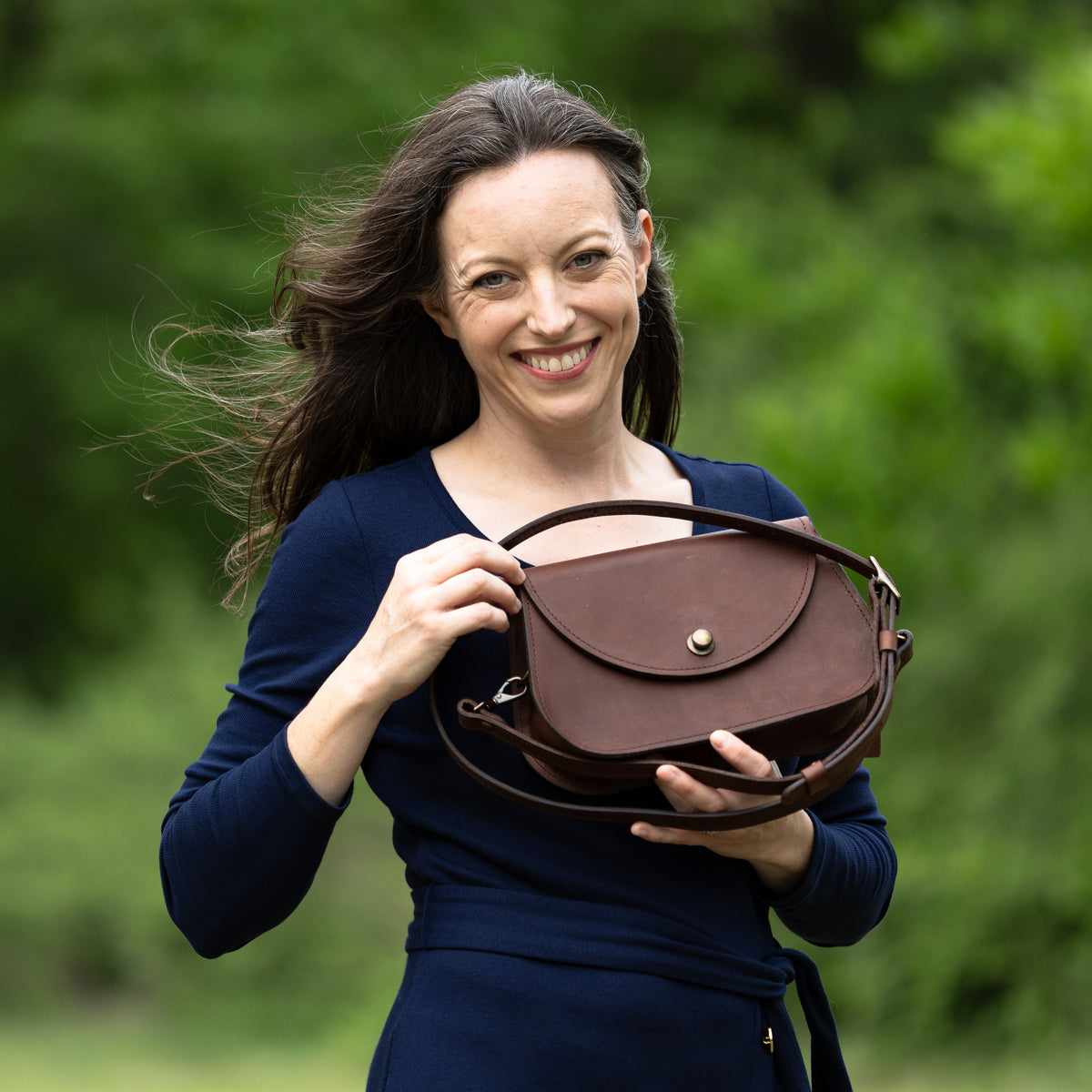 Red Ethel Tote Bag  with russet tan vegetable tanned straps hanging from model's elbow. Model is wearing a blue dress with pink, white and yellow flowers and is leaning against a tan stone wall.