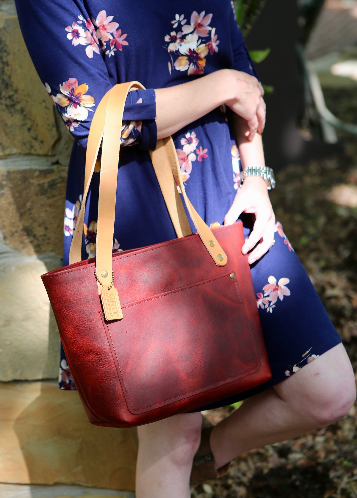 Red Ethel Tote Bag  with russet tan vegetable tanned straps hanging from model's elbow. Model is wearing a blue dress with pink, white and yellow flowers and is leaning against a tan stone wall.